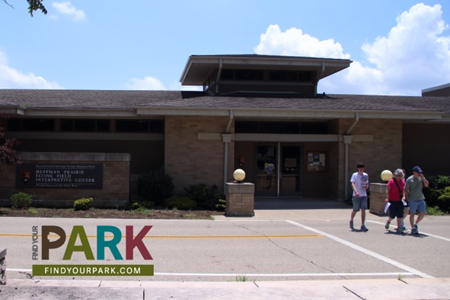 A long, wide one-story building with three people walking away from it in the crosswalk of a road.