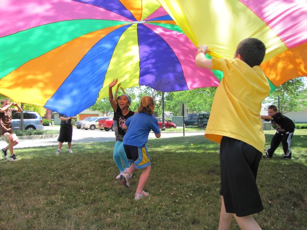 A group of kids hold up a multi-colored sheet while two other kids run underneath it. A group of kids hold up a multi-colored sheet while two other kids run underneath it.