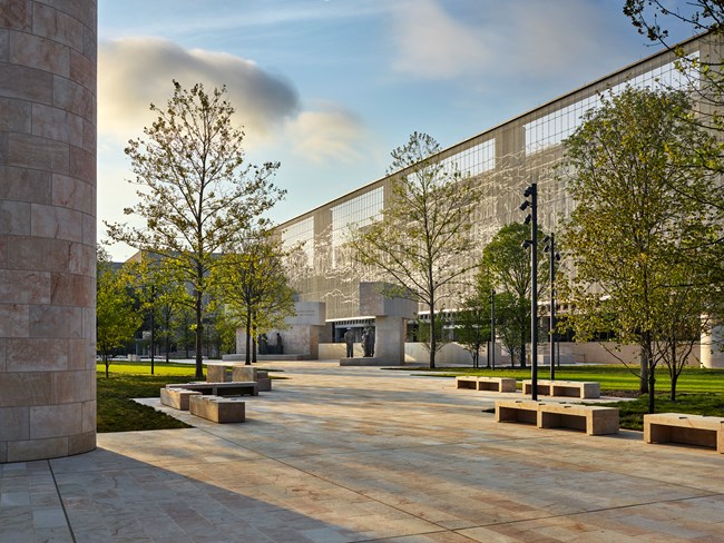 The surface of the memorial is white granite. A large, tile art piece is displayed hanging in the back. A large column is seen to  the left. Two sets of statues stand in the middle toward to background.