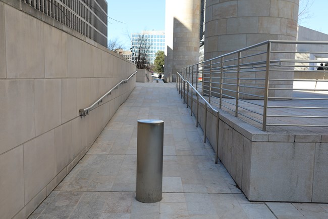 A ramp is positioned between the memorial and the U.S. Department of Education building. A bollard is positioned at the bottom and in the middle of the ramp. A handrail is positioned on the left.