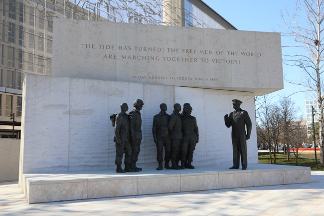 A memorial of one statue speaking to five statues of soldiers is positioned on a raised platform. Engraved words are positioned above the statues on a stone wall.