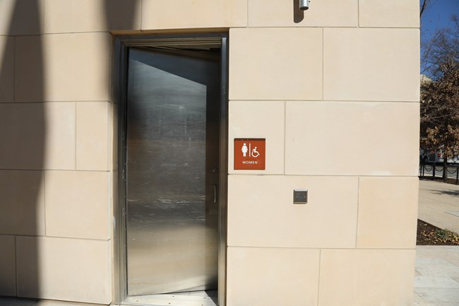 A tall, metal door is positioned to the right of a sign that reads, "women". To the right of the sign is a wheelchair-accessible door button. The door also had a handle for visitors to push open.