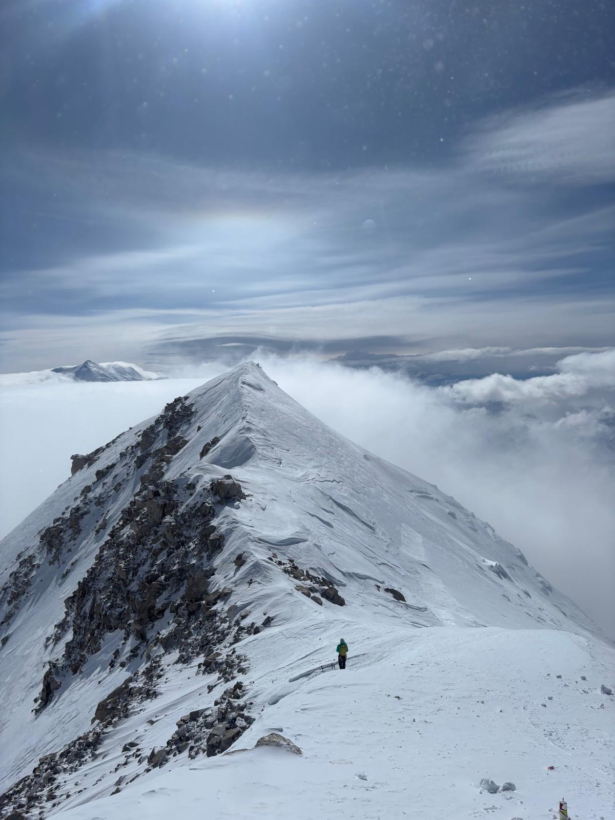 1 person stands near the rise of a snow-covered ridge that looks out on to a layer of white clouds and blue sky