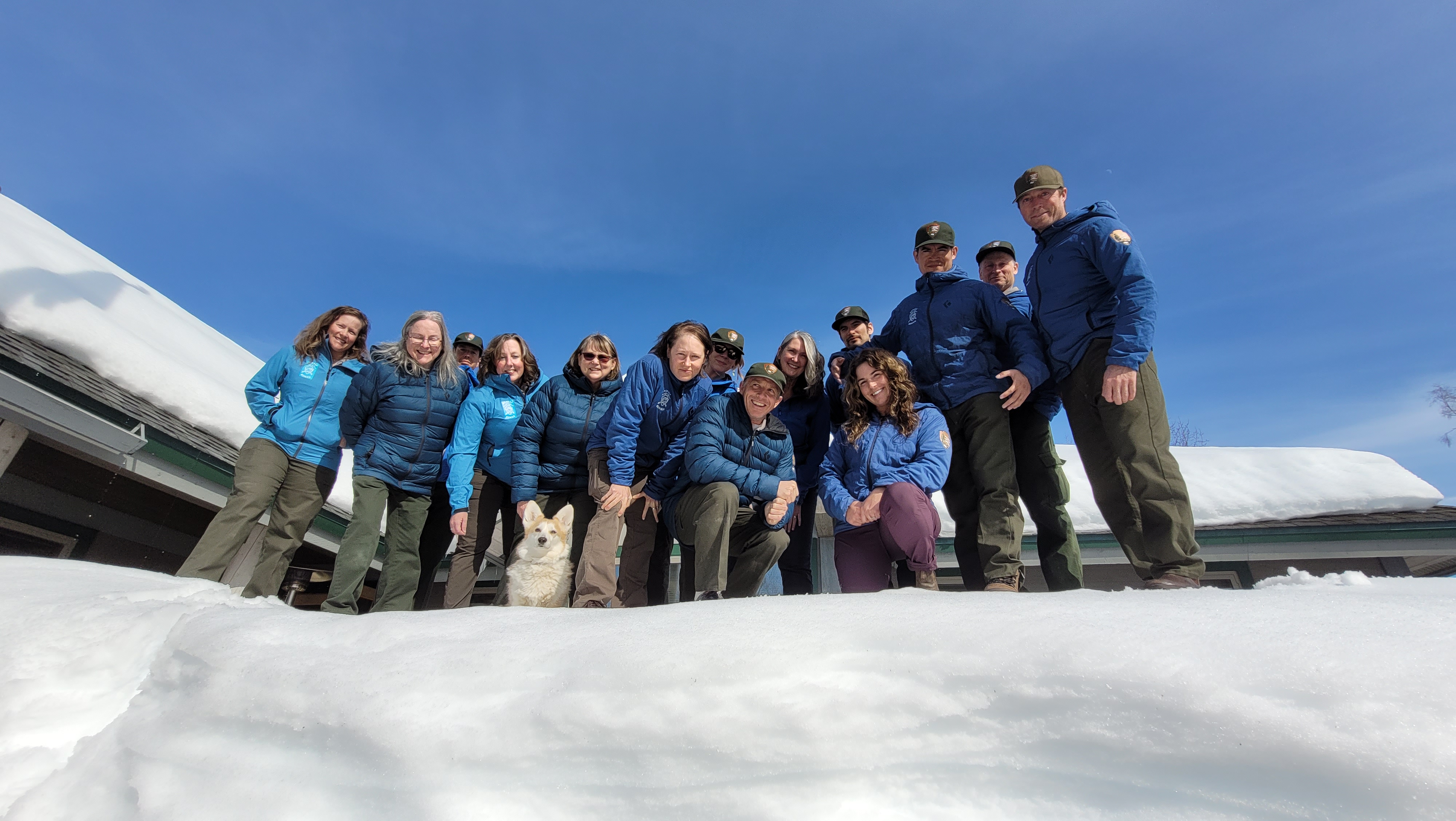 A work team looks down at the camera from a high snow bank outside of a ranger station