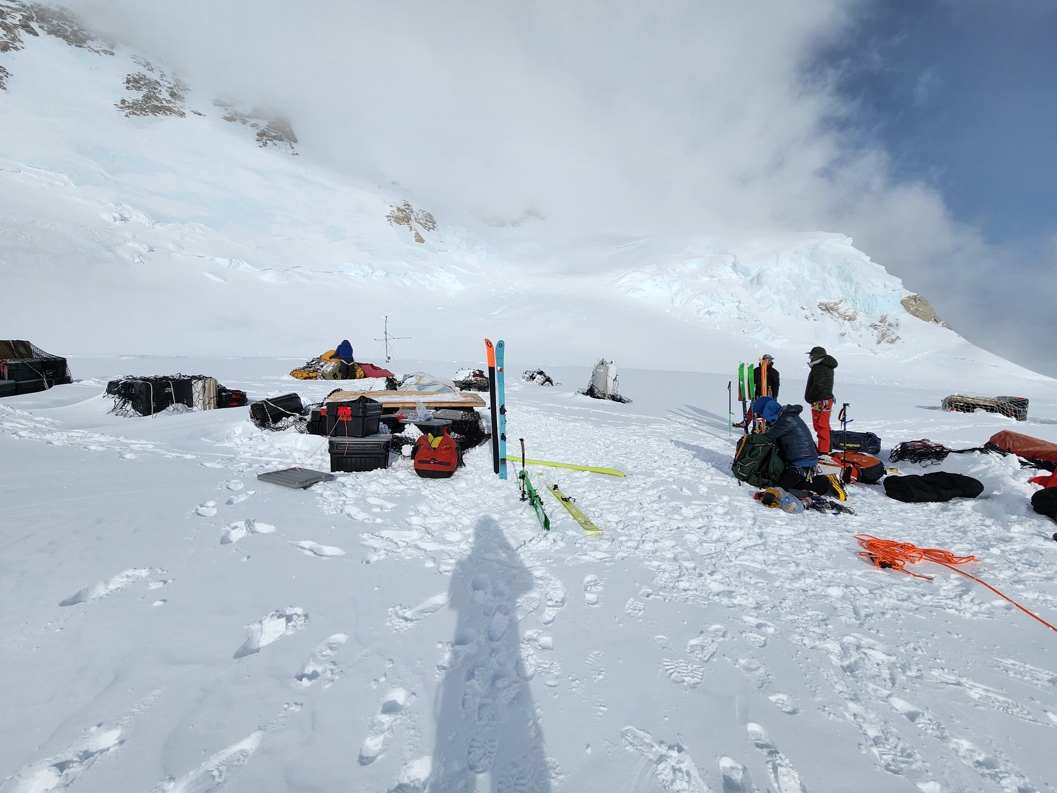 A snowy glacier covered with random piles of plastic storage boxes, duffel bags, and skis