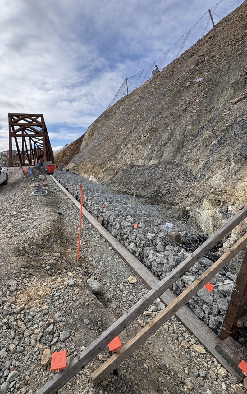 A row of wire cages filled with rocks sits along the shoulder of the road at the base of a steep slope. Behind the constructino site, the Pretty Rocks bridge is visible.