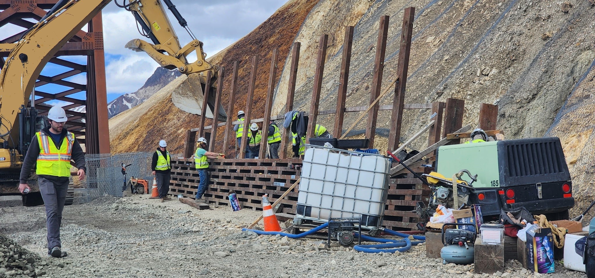 A wooden fence is partially constructed at the base of a steep slope. Several construction workers work on the fence.