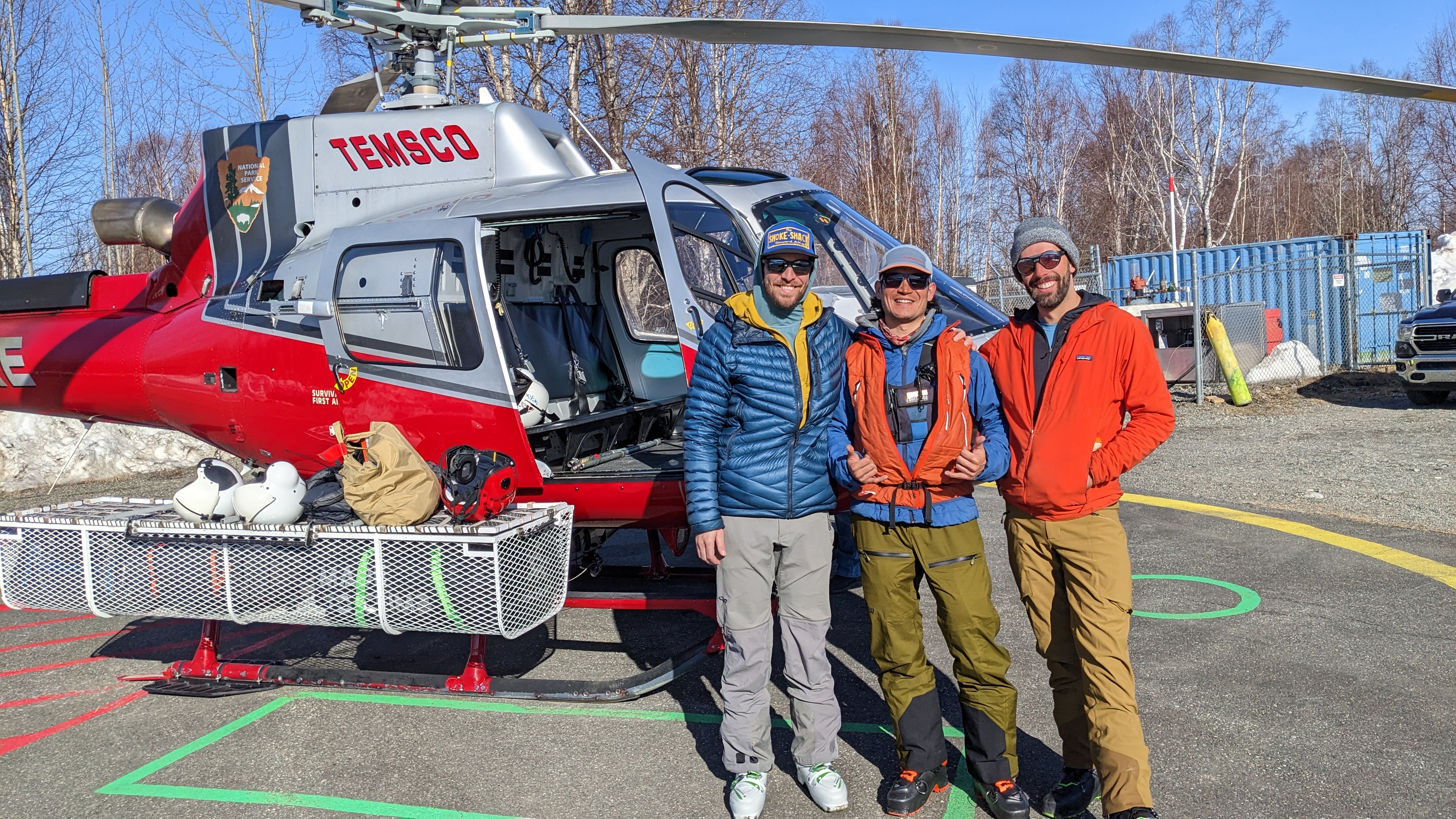 Three men in sunglasses and outdoor gear stand outside a grey and red helicopter