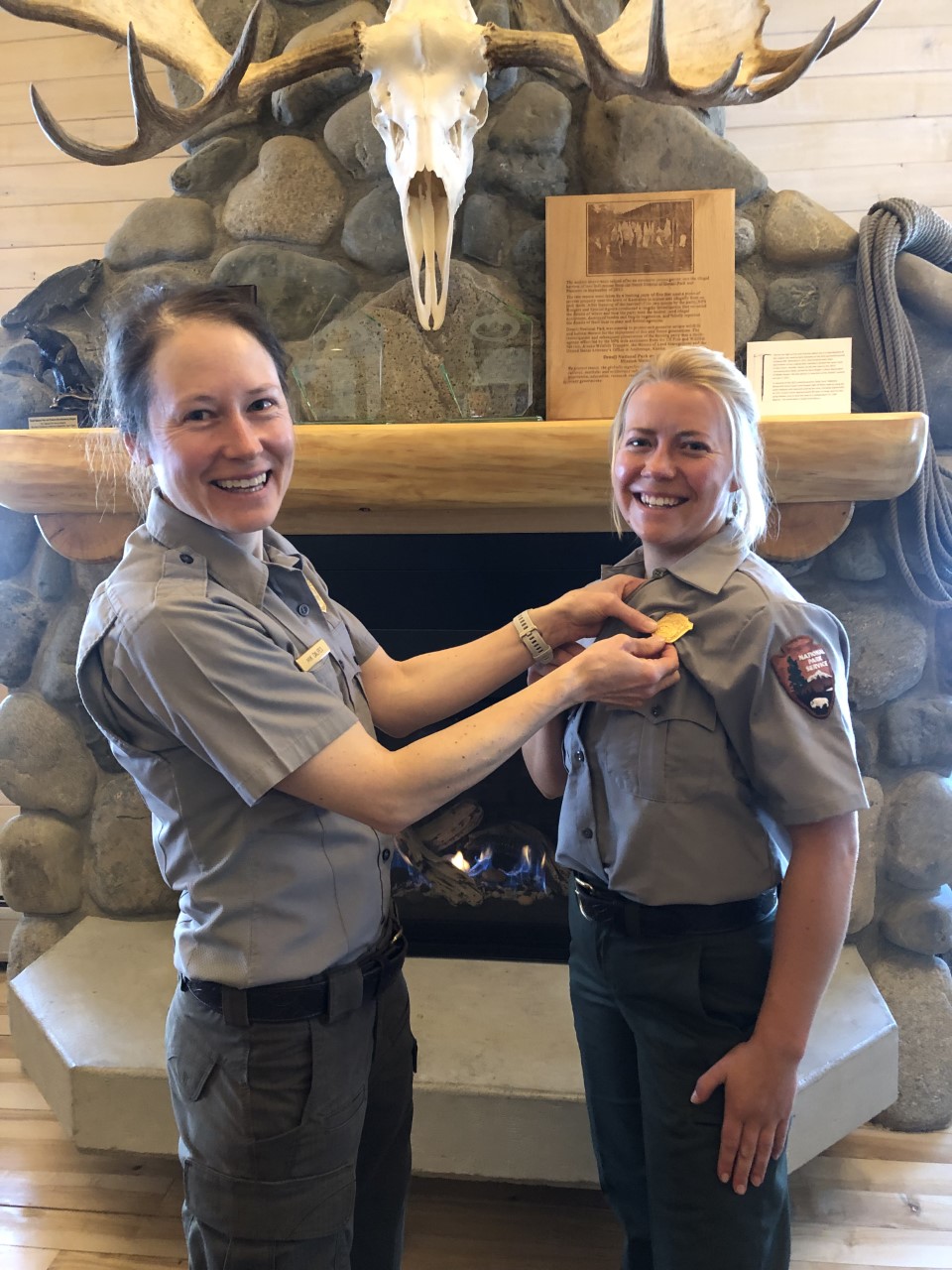 A woman in park ranger uniform pins a gold badge on another woman in a ranger uniform in front of a stone fireplace.