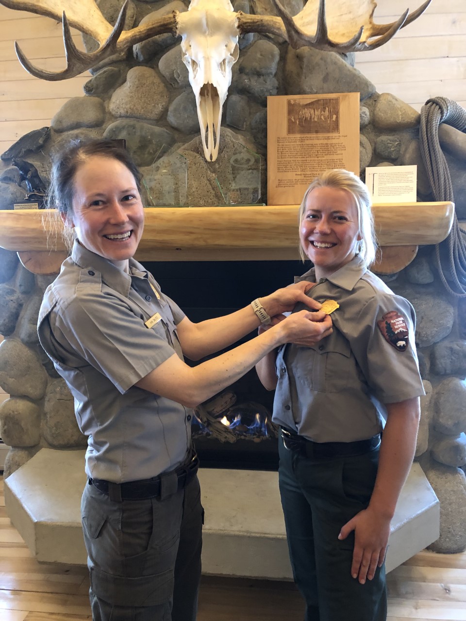 Welcome to new mountaineering ranger Anika Ramey! A woman in park ranger uniform pins a gold badge on another woman in a ranger uniform in front of a stone fireplace.