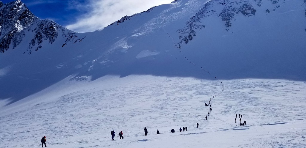 A long line of climbers ascend a mountain ridge