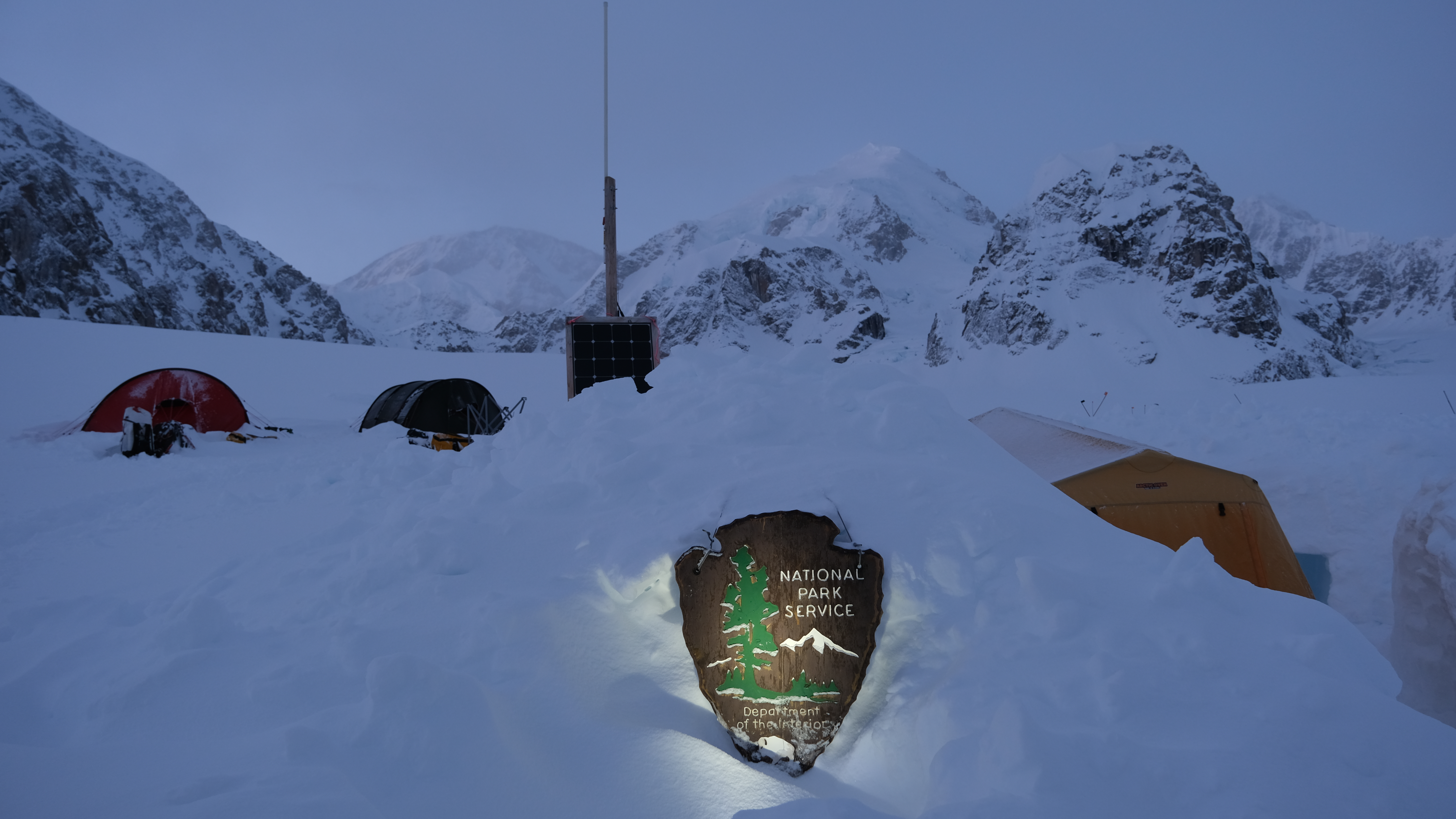 An NPS arrowhead sign is illuminated at a glacier encampment at dusk
