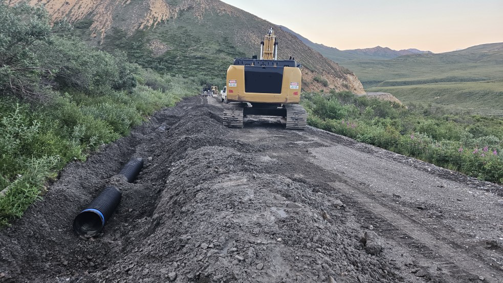 Heavy equipment on a narrow gravel road. On the left shoulder of the road, a piece of a culvert piece lies in a ditch.