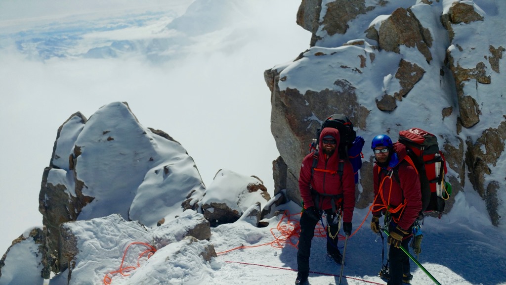 Two mountaineers with heavy backpacks stand near a rocky ledge
