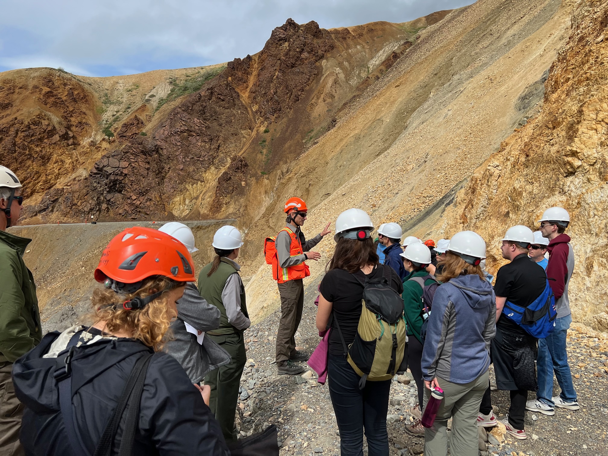 CODEL 6 A group of about 15 people wearing protective hard hats stands at the edge of the Pretty Rocks landslide, where the gravel road bed drops away and slumps down a rocky mountainside. A few park staff are in uniform and one man speaks to the whole group.