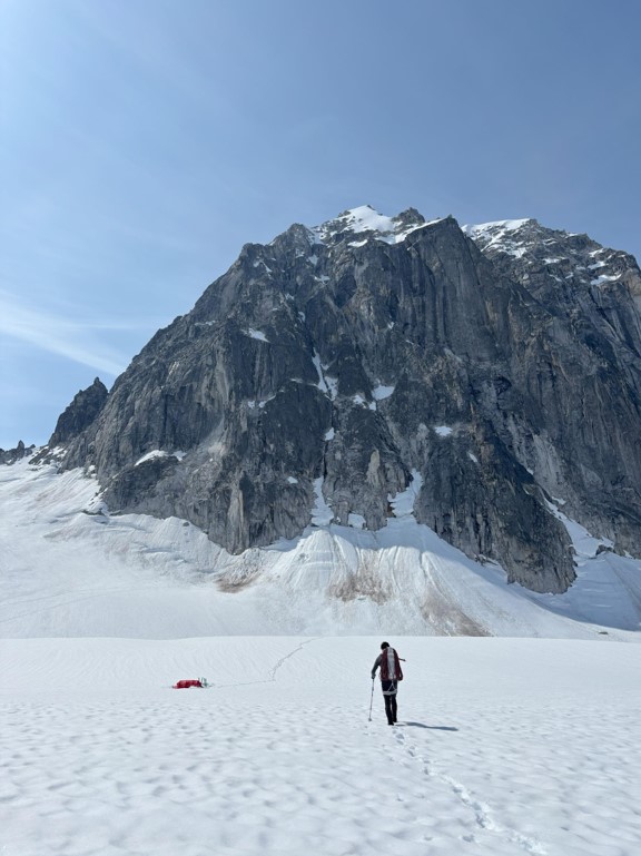 Camping below the Royal Tower with the route Ranger walks across a snowy field toward a red tent with mountain peaks in the background