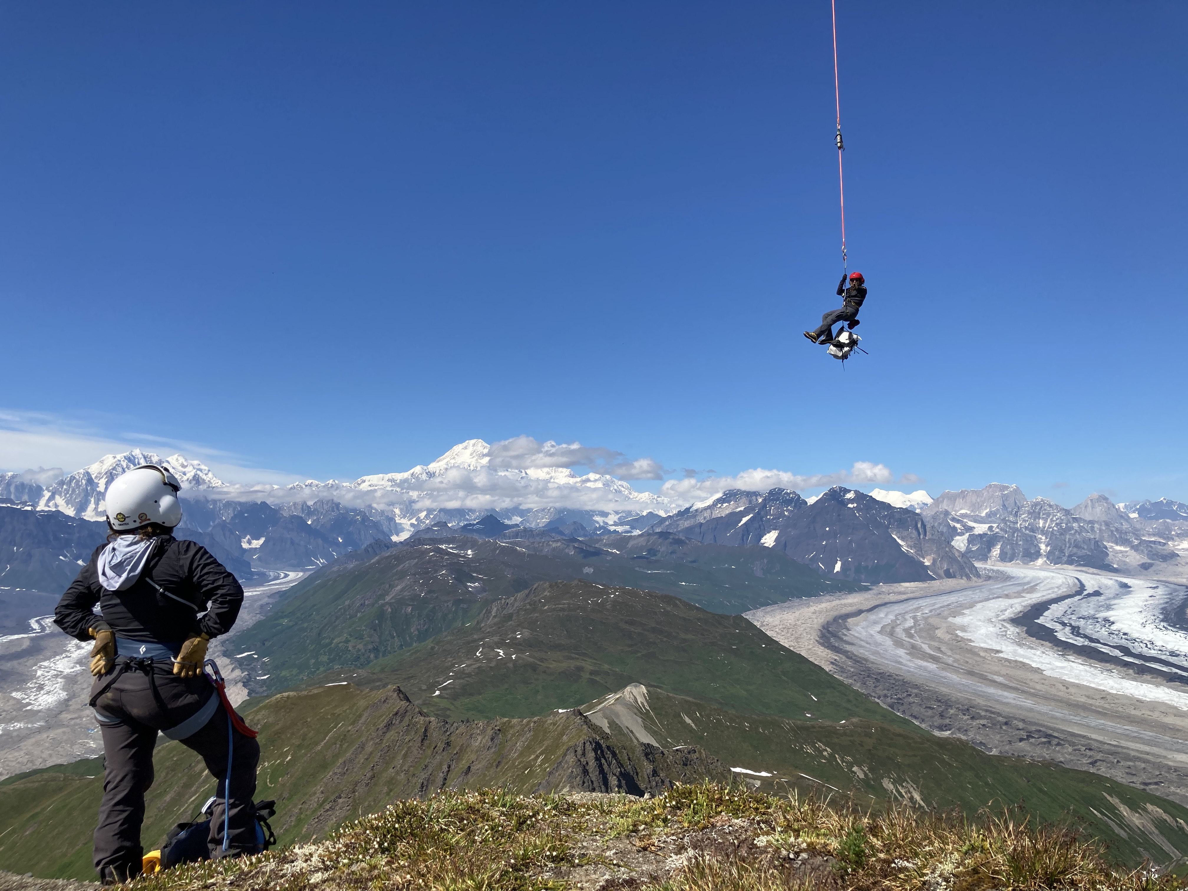 A ranger on a green hillside peers out at a fellow ranger hanging mid-air from the end of a helicopter ropeline, with snowy Denali in the distance 