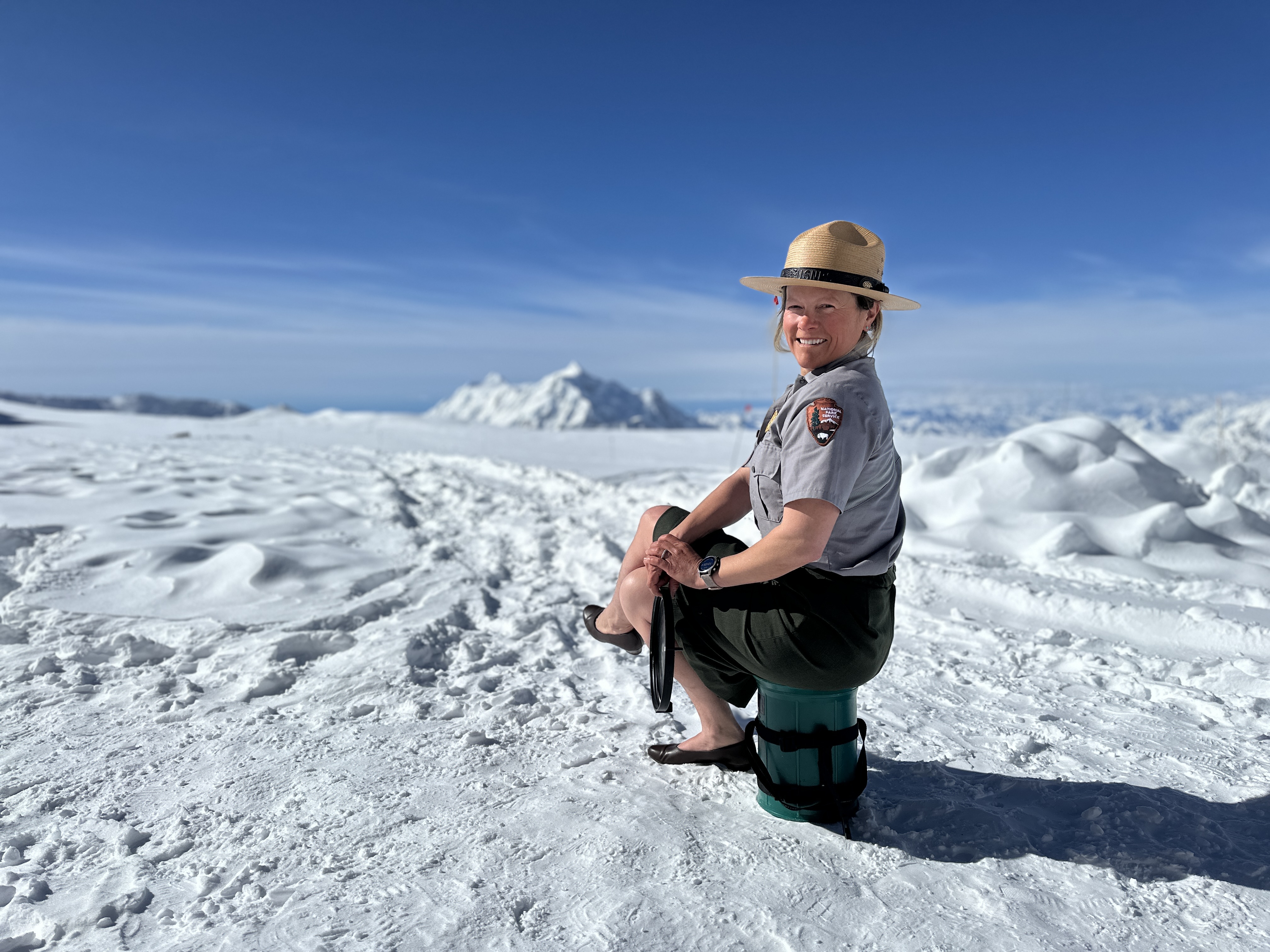 Ranger Chrissie takes in the view A smiling park ranger in an NPS flat hat, skirt, and pumps sits atop a green Clean Mountan Can, with a snow covered peak in the distance