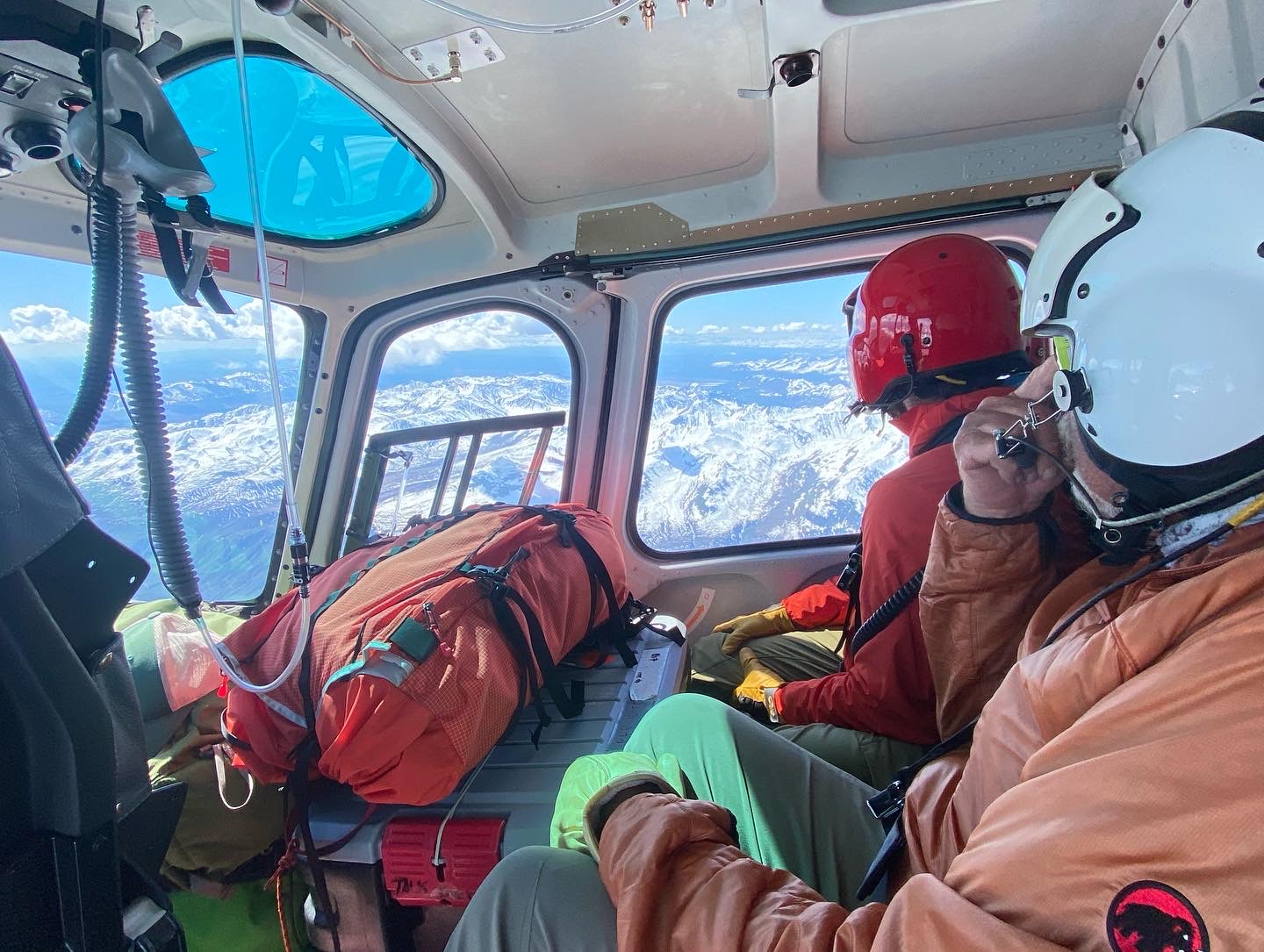 Two climbers look out a helicopter window to mountains below