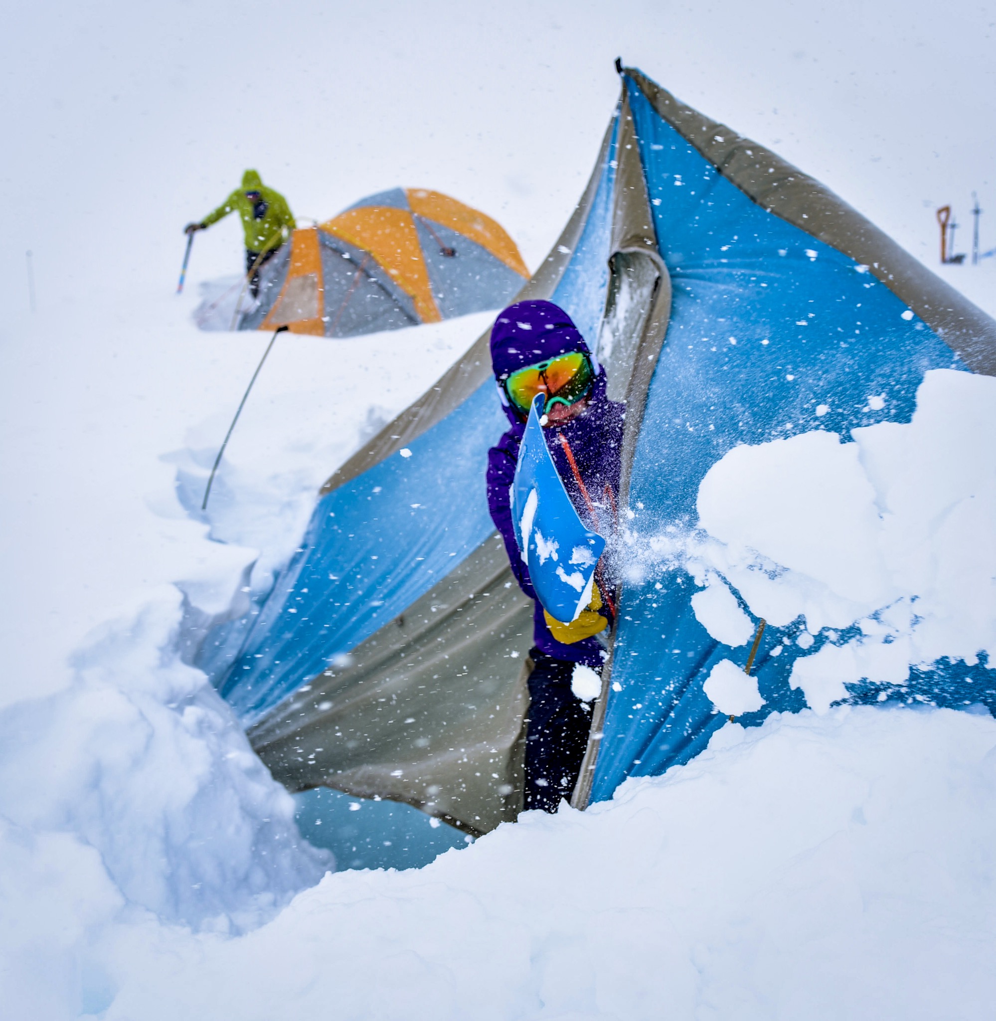 A technicolor-clad climber shovels snow from their heavily-laden tent