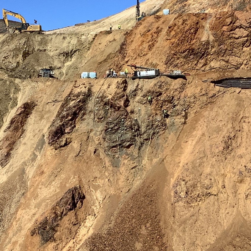 Drilling Blast Holes Two people work on the side of a cliff supported by ropes with construction equipment above them.