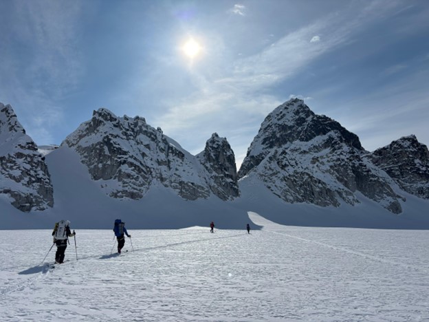 Four skiers in a line across a snowy landscape with mountains and a blue sunny sky in the background