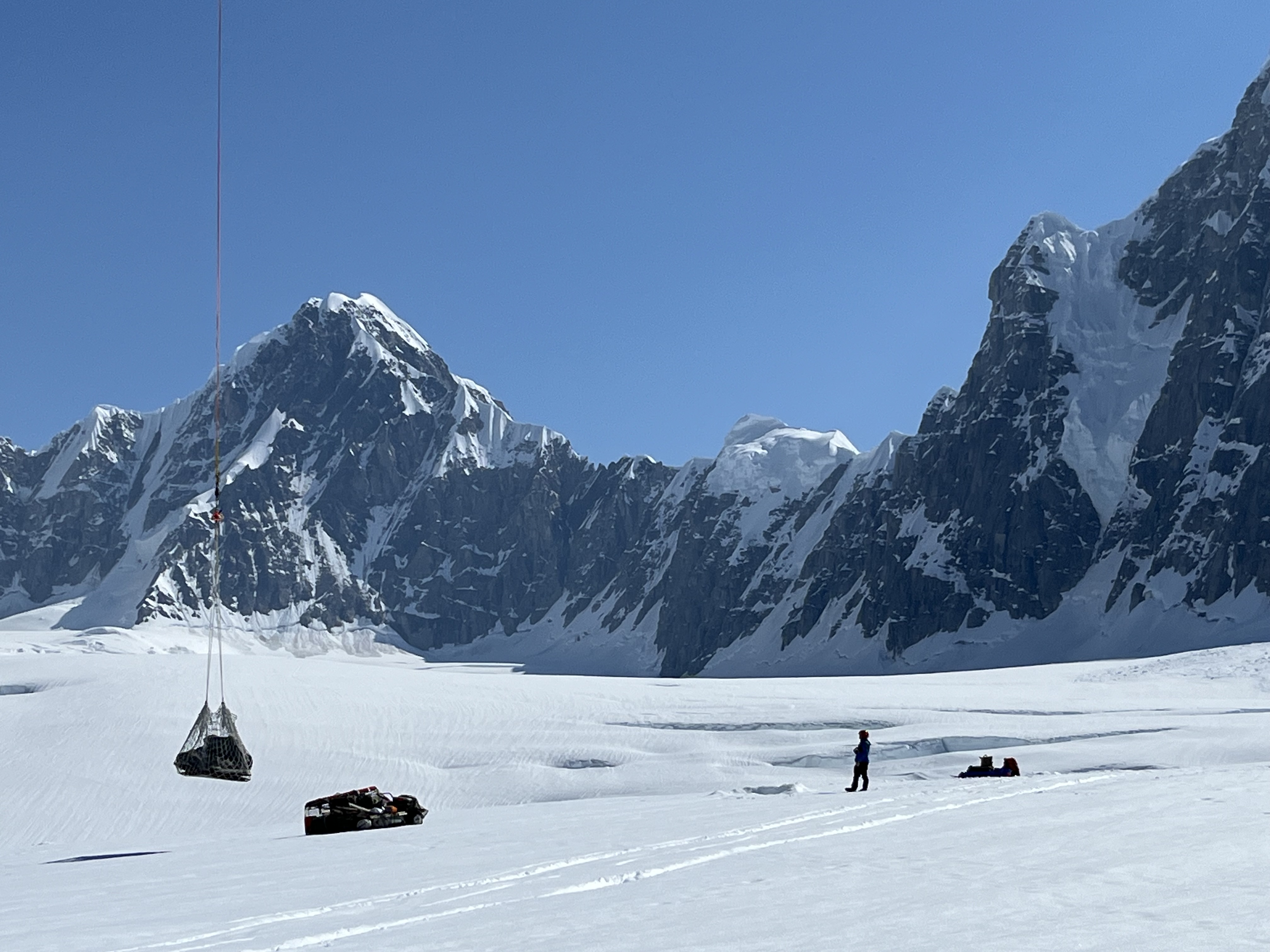 Sling-loading 14,200 foot camp gear A silhouette of a ranger standing on a glacier observing a net load of equipment suspended at the end of a long rope line.
