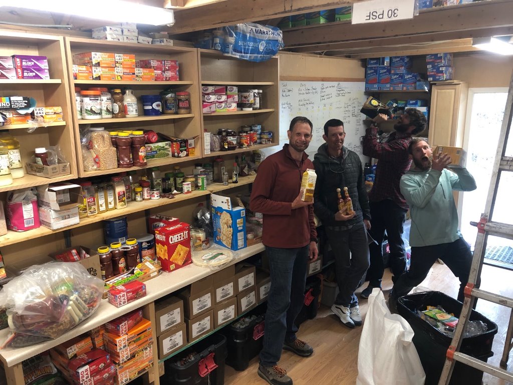 Four men stand in a well-stocked food storage room