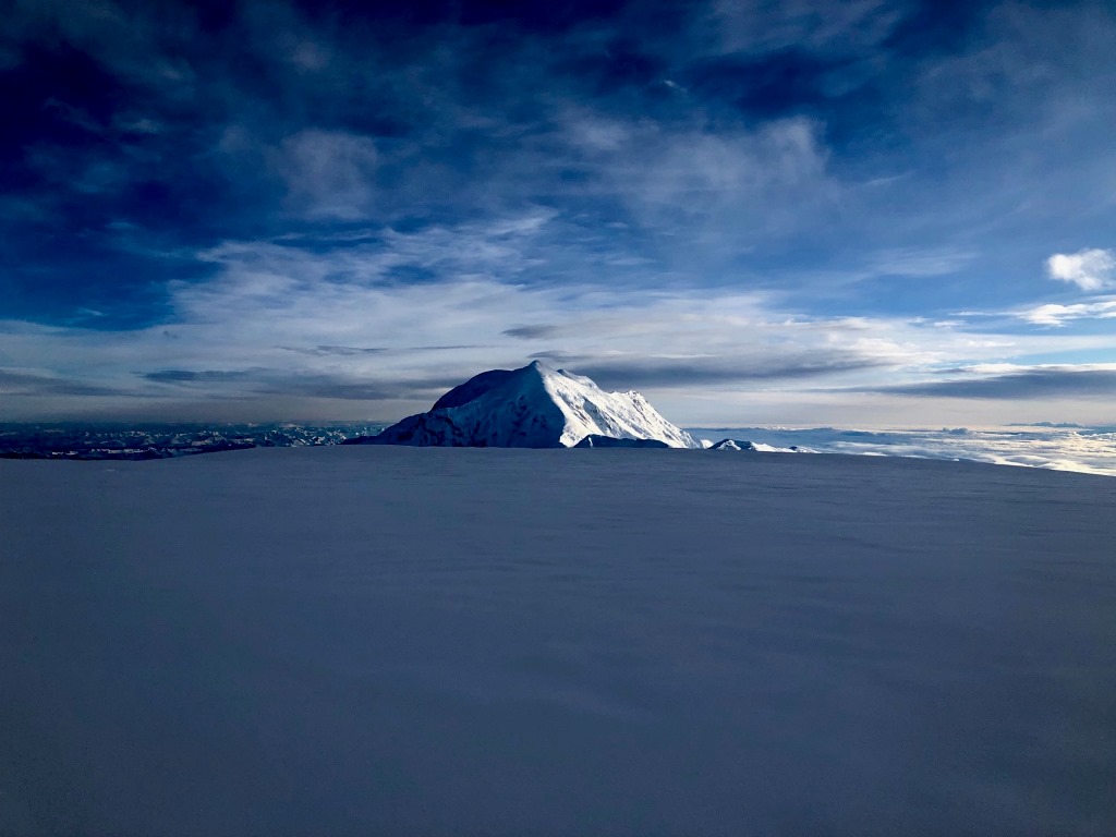 A jagged mountain peak, half in shadow and half in shade, appears above a glacier horizon and a sky with wispy clouds