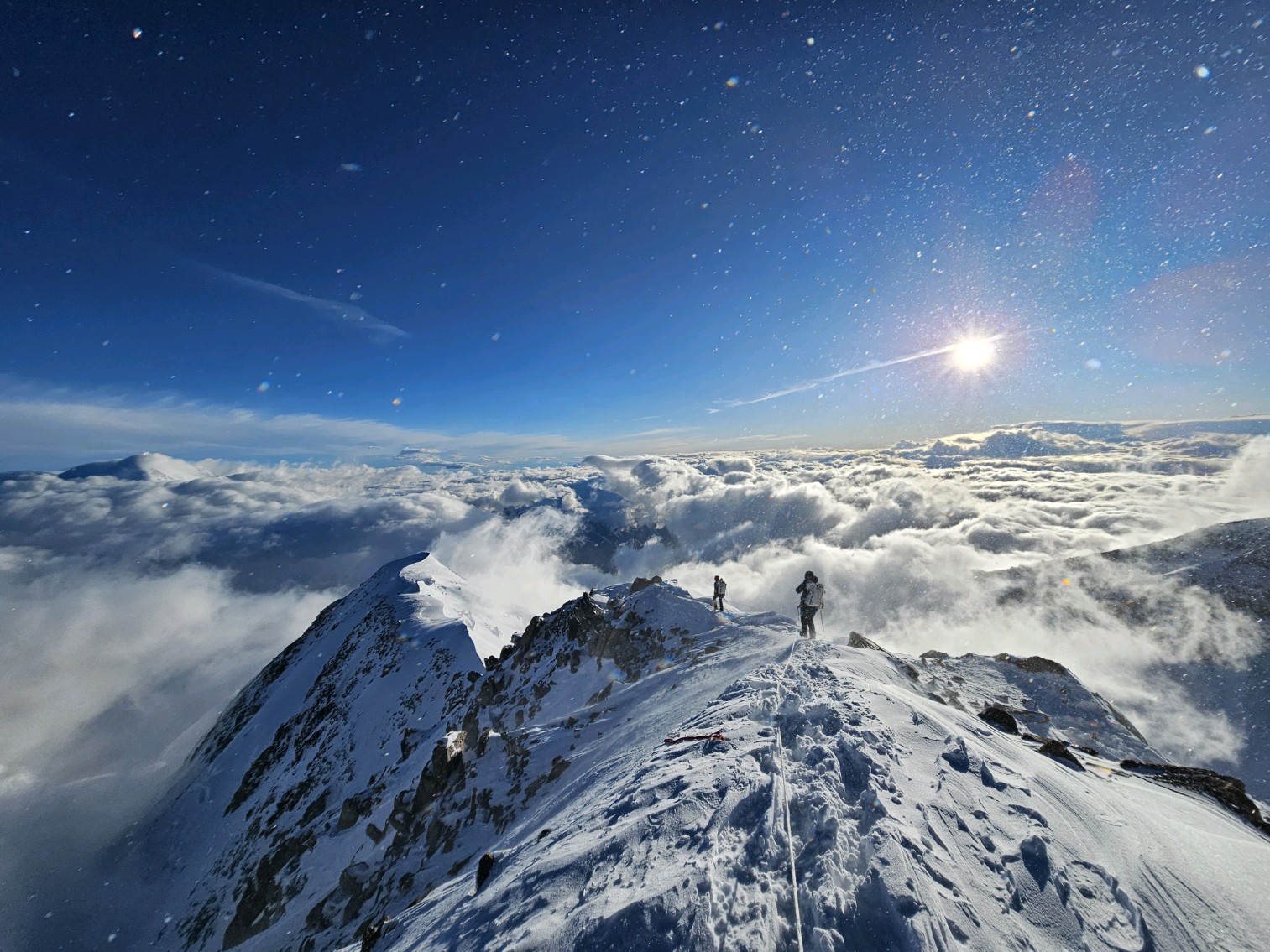 View looking down as two climbers descend a narrow rock and snow ridge with a setting sun in the distance