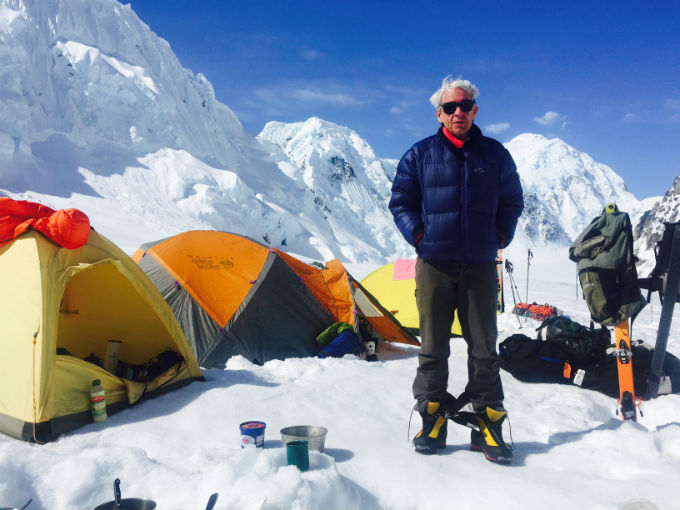 Colorful photo of a climber standing in a tent camp, surrounded by blue skies and white mountain peaks