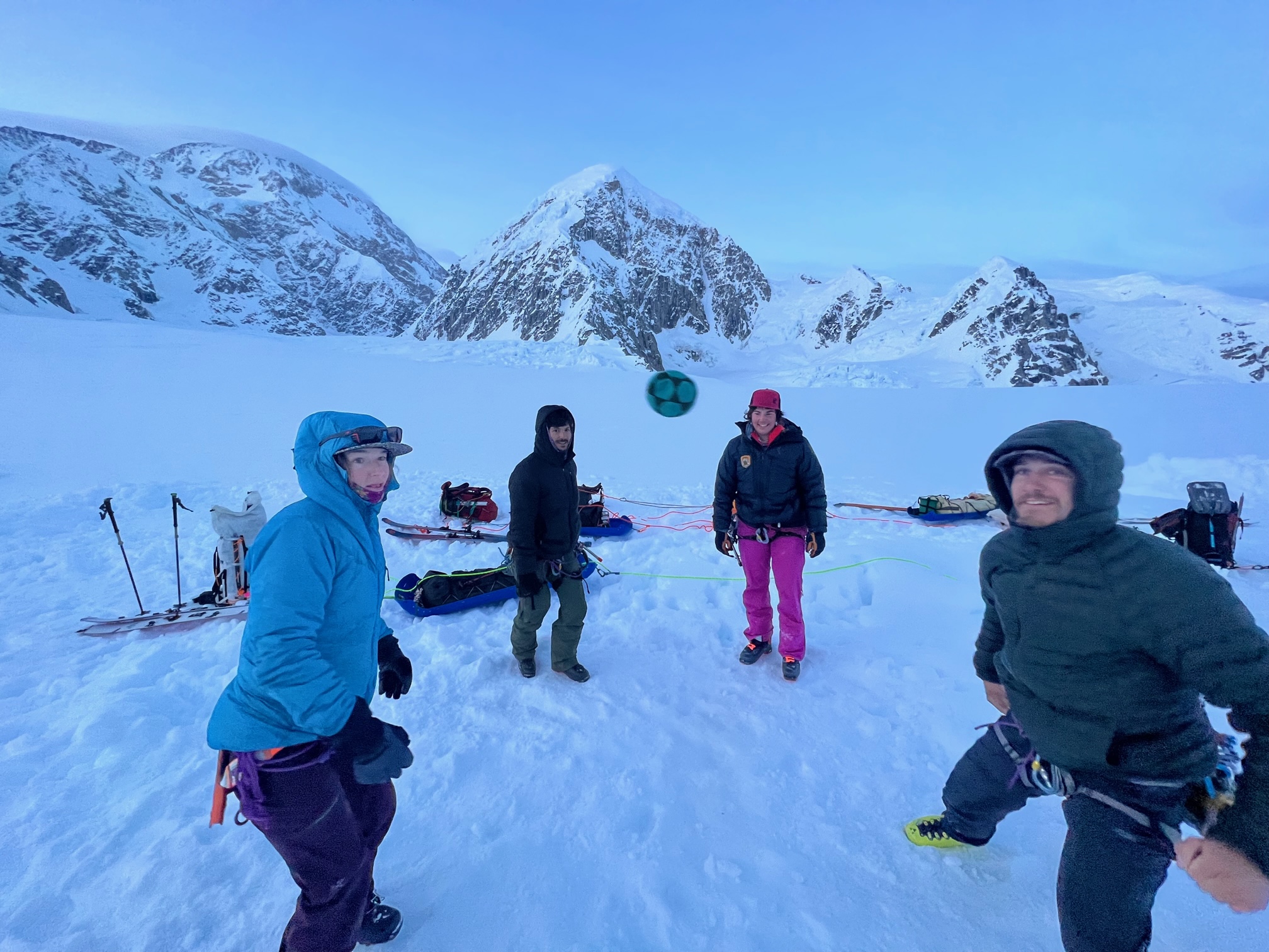 Four climbers stand on a glacier look upon a hackysack in mid-air