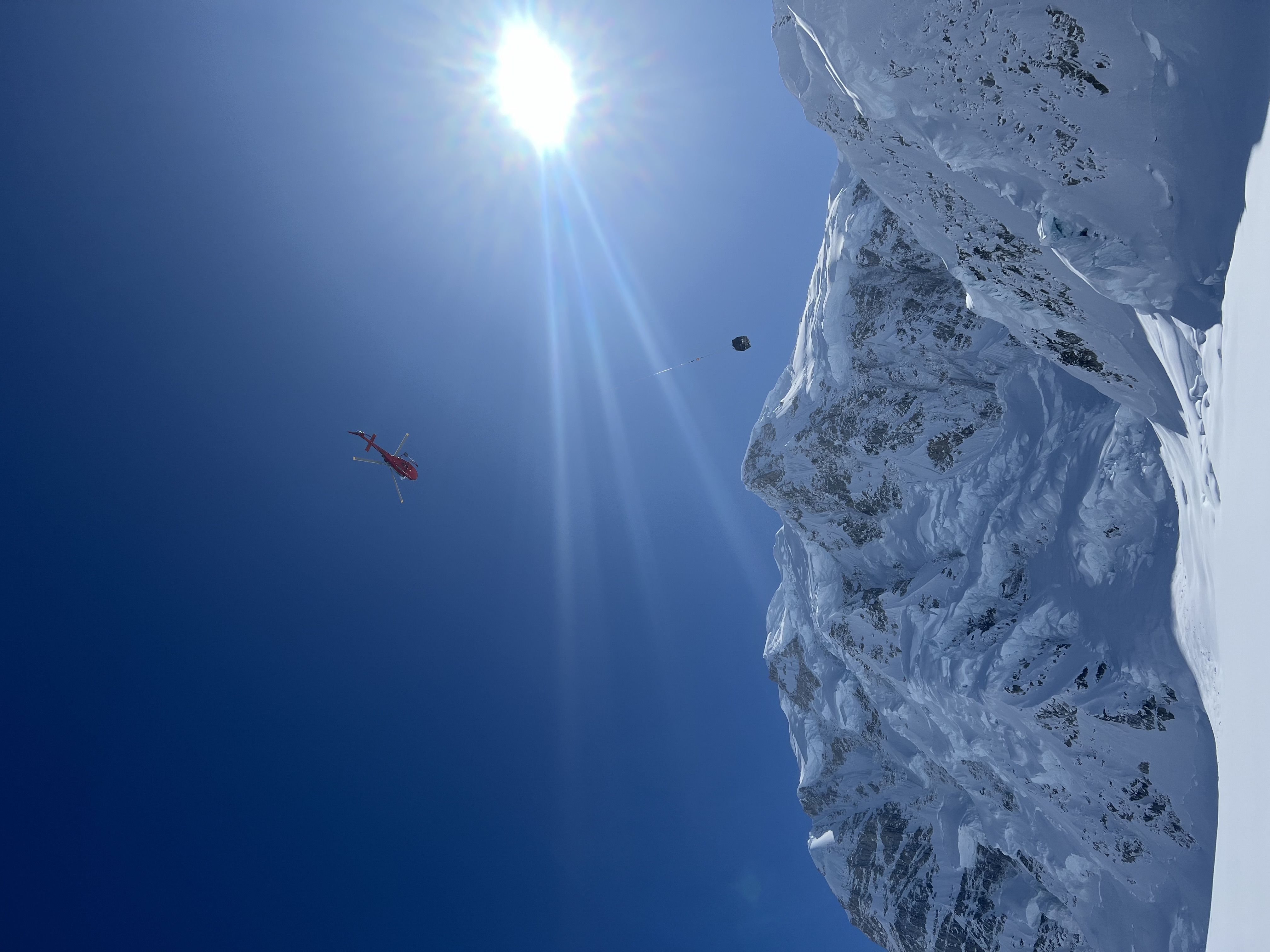 Hauling camp loads to 14,200 feet A helicopter flies past a snow-covered mountain hauling a cargo netload at the end of long rope line