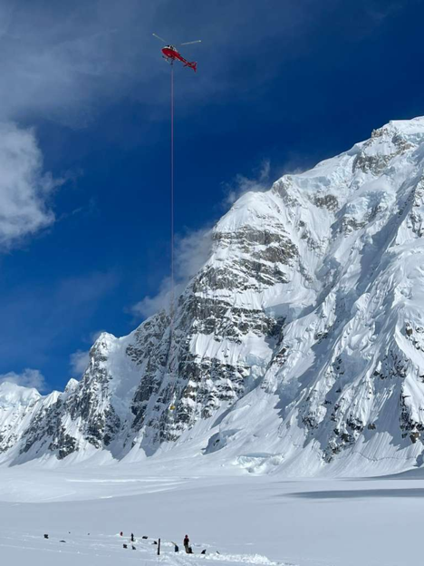 Helicopter 1 A red helicopter hovers high above a snowy landscape with a rope lowered to a ranger standing below it in front of a large mountain peak and blue sky.
