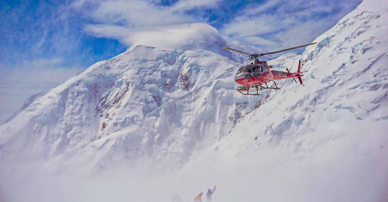 A red helicopter hovers above two climbers on the glacier below