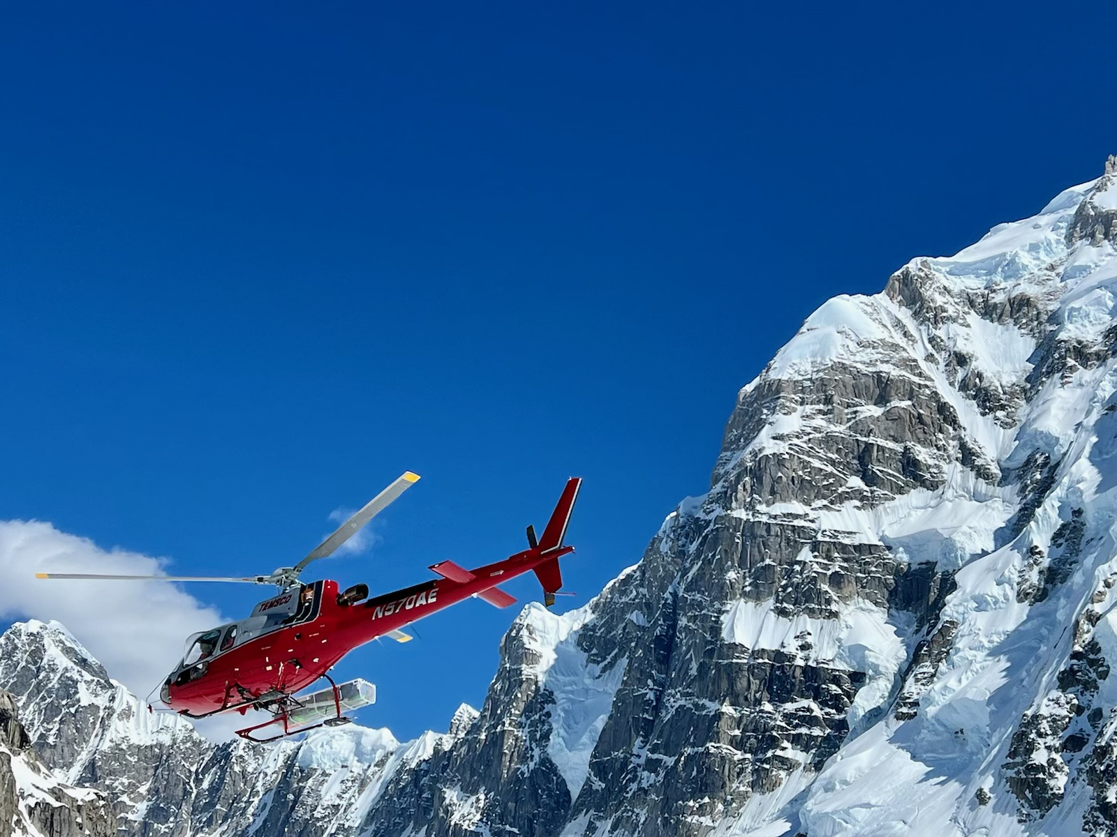 A helicopter flies in bright blue skies along a rock and snow ridge.