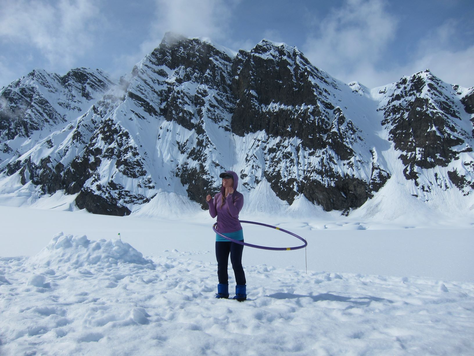 A woman twirls a hula hoop, standing on a glacier with granite peaks in the background