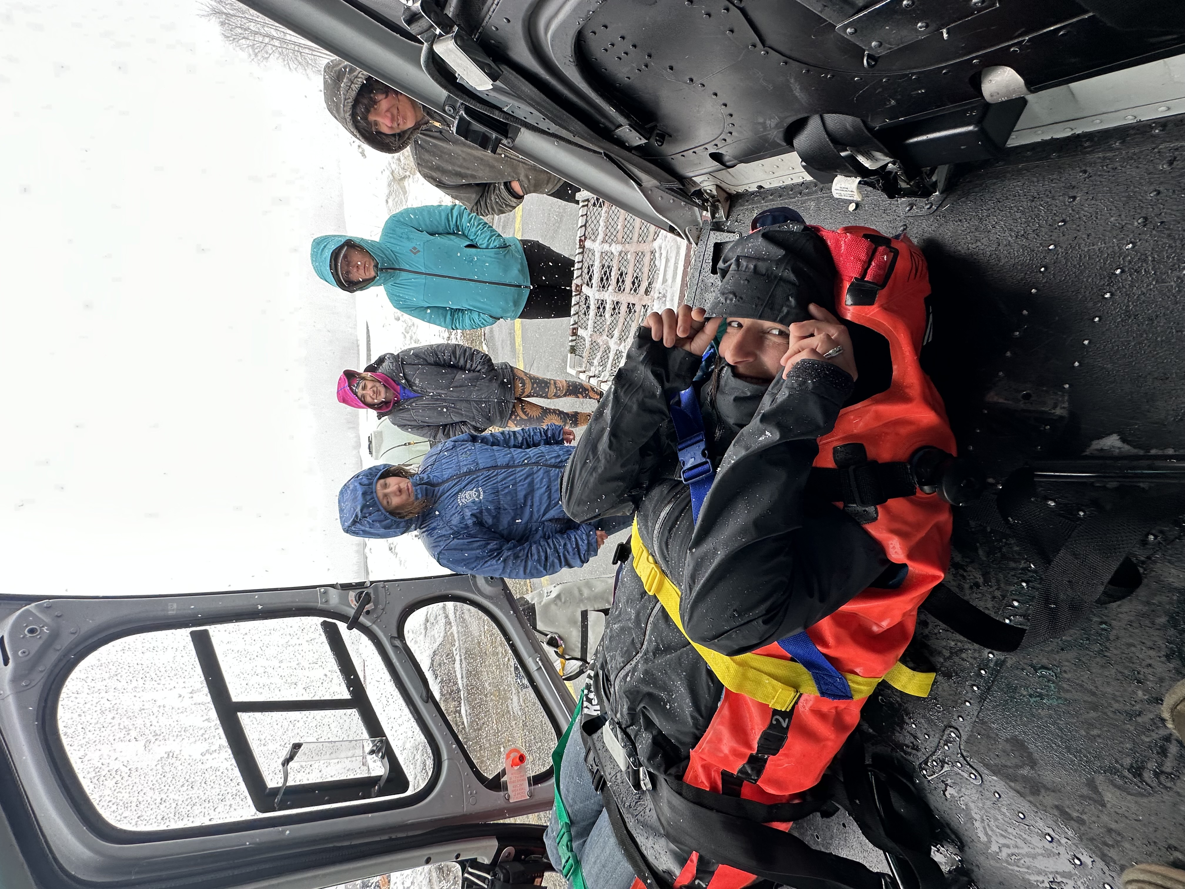 A patient buckled into a stretcher on the floor of a helicopter smiles up at the camera. Four hooded women observe from outside the helicopter, standing in a wet snowstorm. 