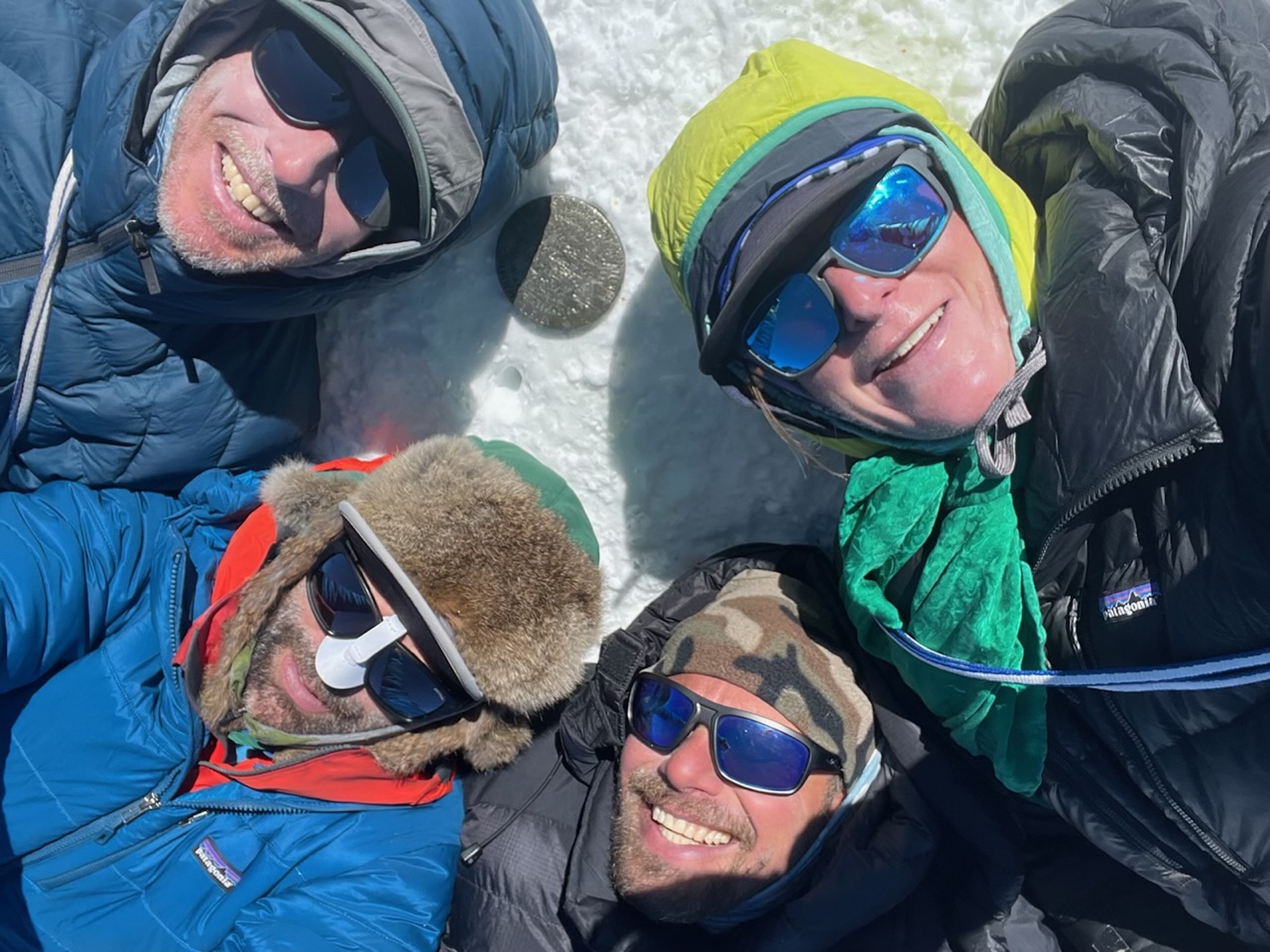 Four smiling faces laying in the now surrounding a USGS summit marker