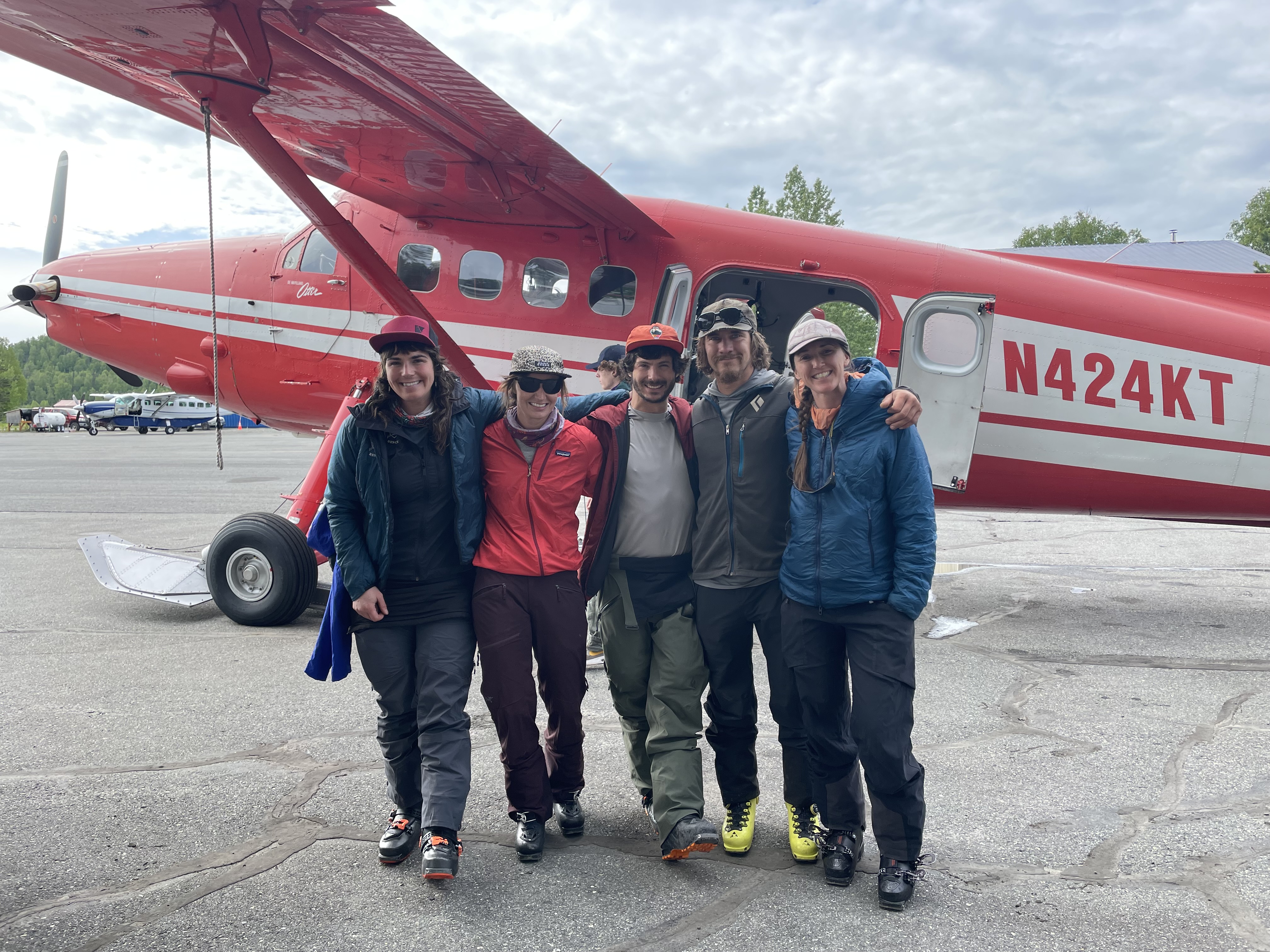 Five climbers link arms in front of a small red aircraft.