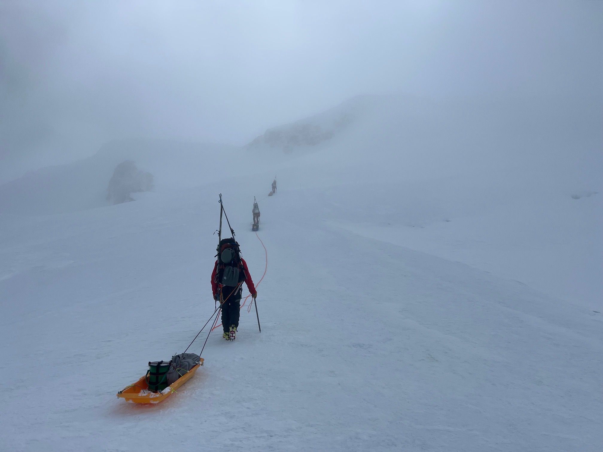 A roped team of climbers pulling sleds moves along a snowy trail in flat light and thin cloud cover