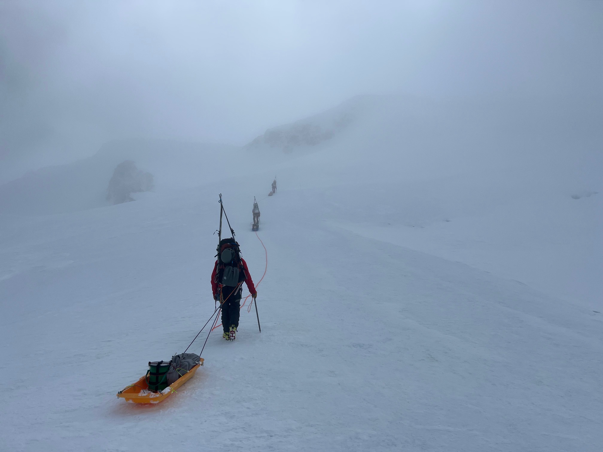 In the mist A roped team of climbers pulling sleds moves along a snowy trail in flat light and thin cloud cover