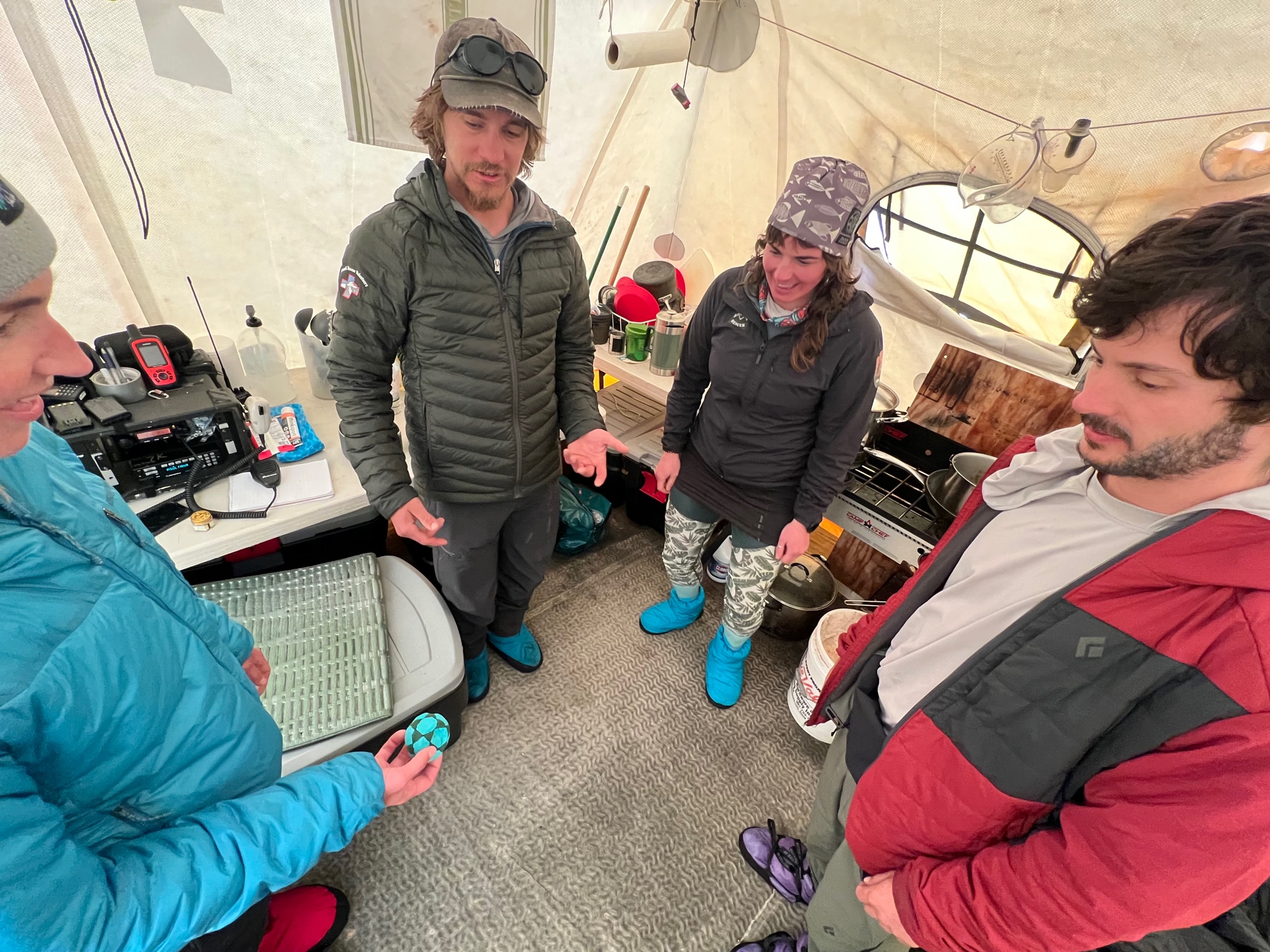 Four climbers standing inside a tent stare down at a hackysack