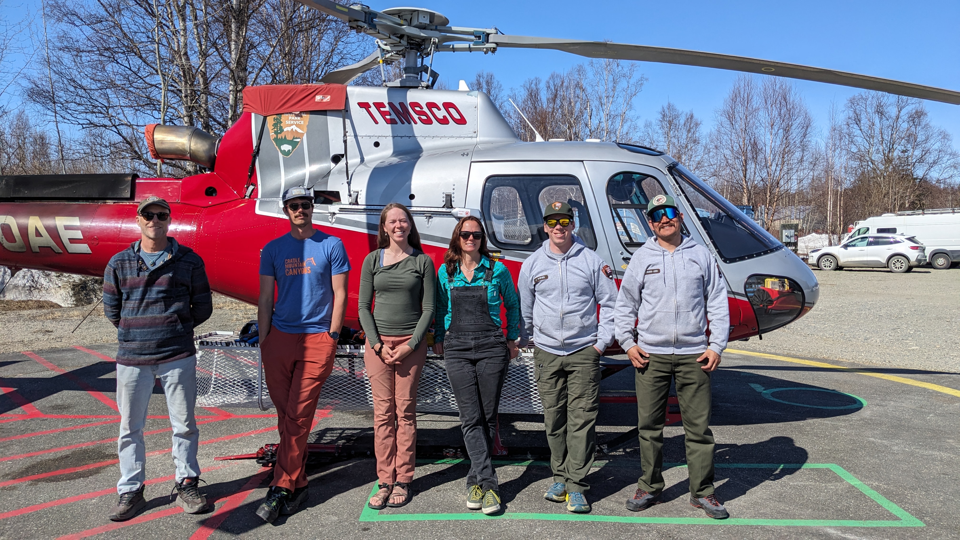 Six team members in street clothes stand next to grey and red helocopter