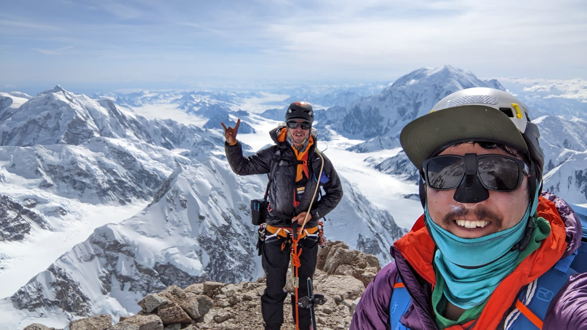 Two men in bright climbing gear, helmets, and sunglasses post on a rock ledge