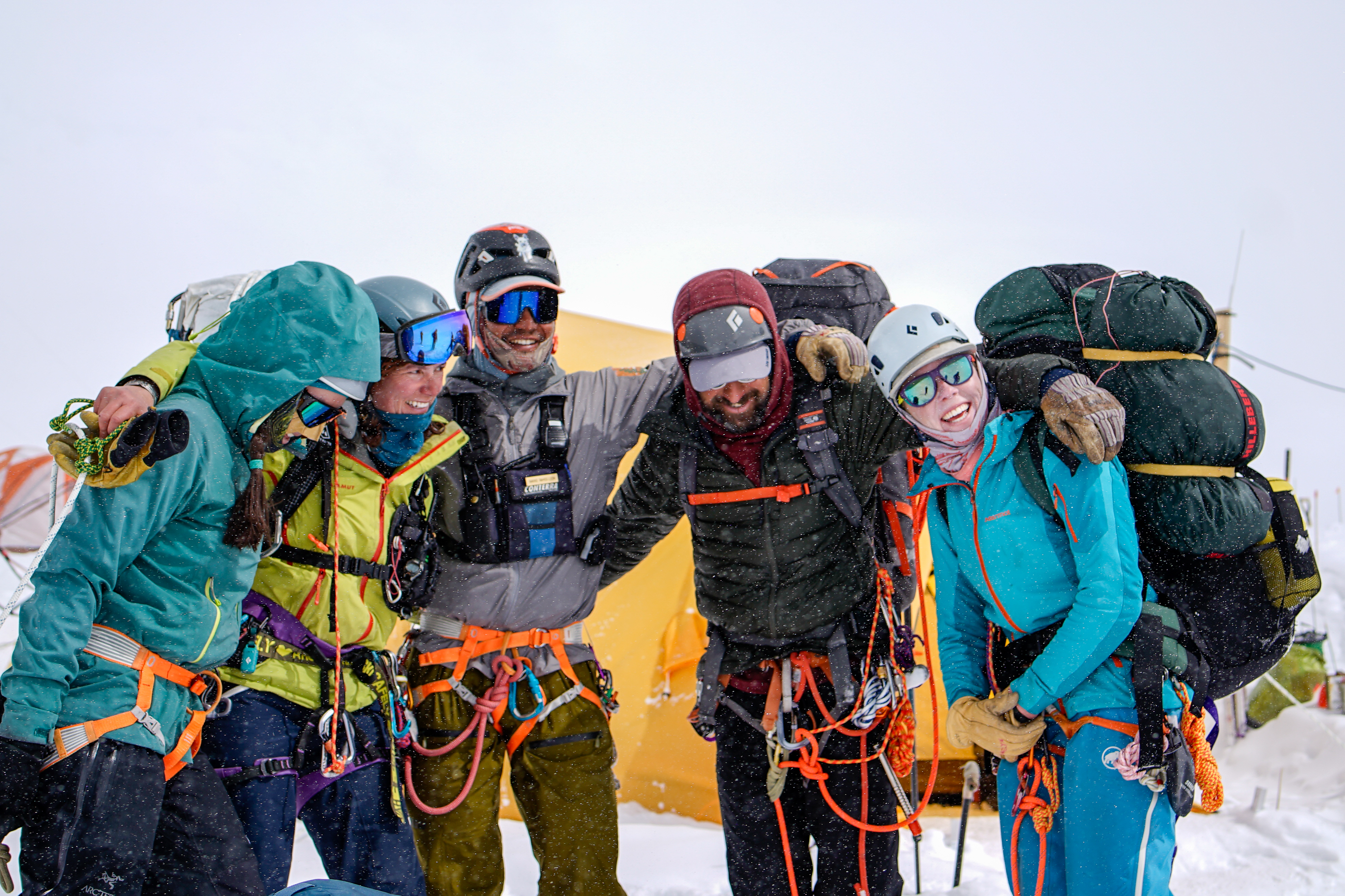 Five helmeted and harnessed climbers carrying big backpacks pause for a photo