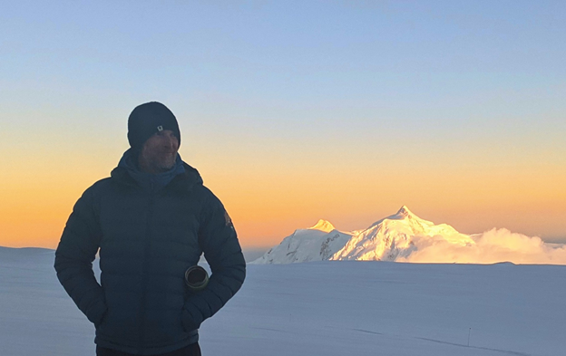Dave's final night at 14,200 feet as a Denali Ranger Shadowed ranger stands on a glacier with alpenglow-bathed peak in background