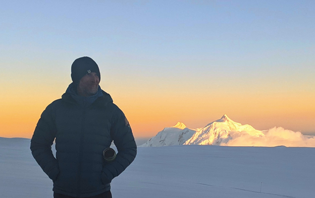 Shadowed ranger stands on a glacier with alpenglow-bathed peak in background