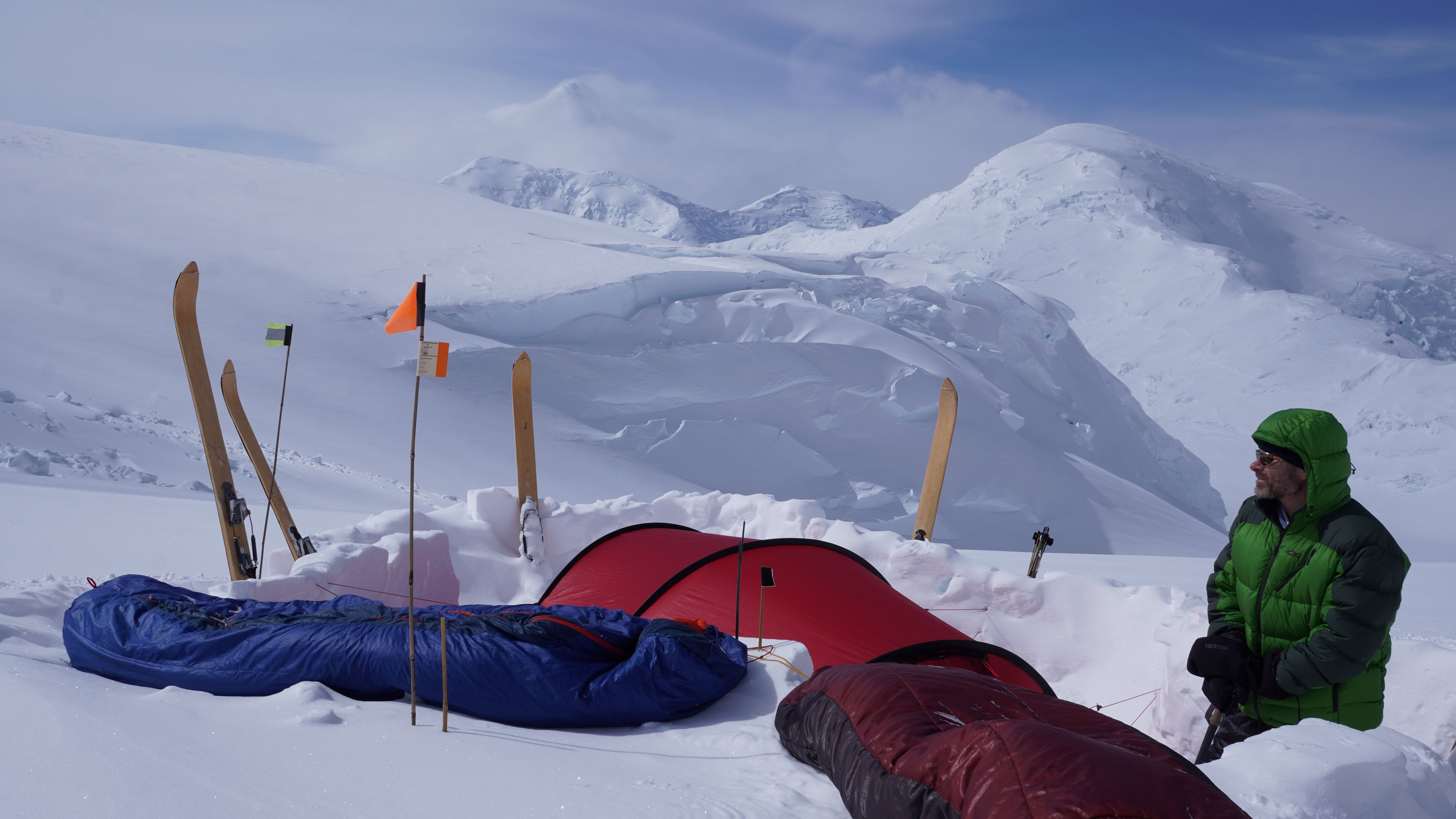 A man in a green puffy coat looks off at the snow capped peaks from his tent encampment