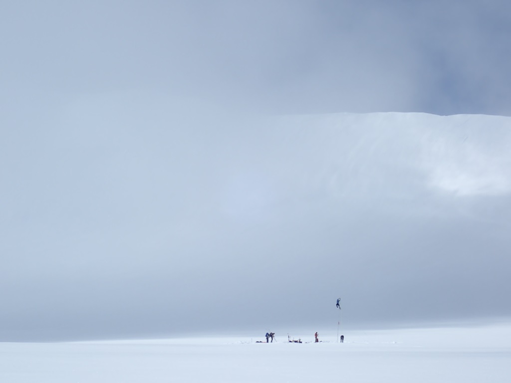 From a distance, a scientist at the top of a skinny weather station pole appears to be flying above the glacier